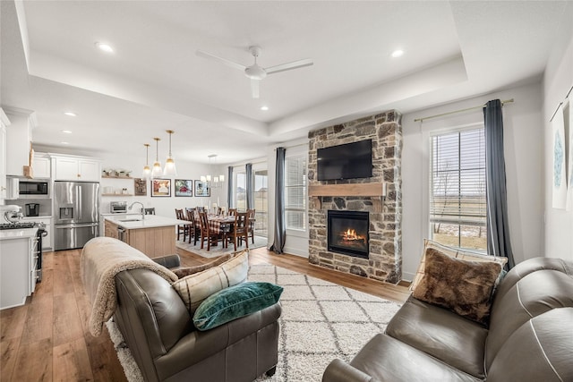 living room featuring ceiling fan with notable chandelier, a fireplace, a tray ceiling, and light wood-type flooring