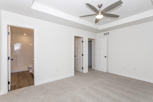 unfurnished bedroom featuring ensuite bathroom, light colored carpet, a textured ceiling, a raised ceiling, and ceiling fan