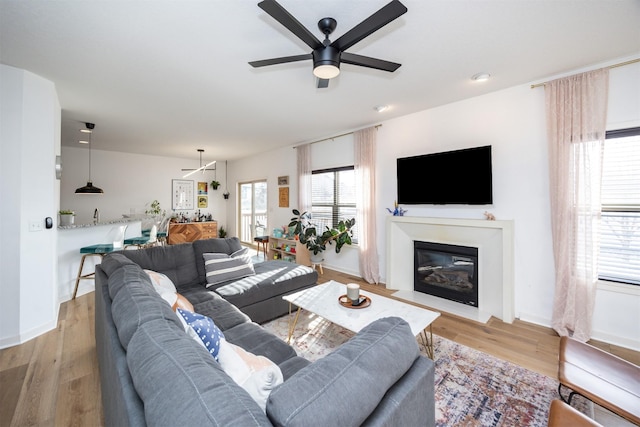 living area featuring a fireplace with flush hearth, light wood-style flooring, baseboards, and ceiling fan