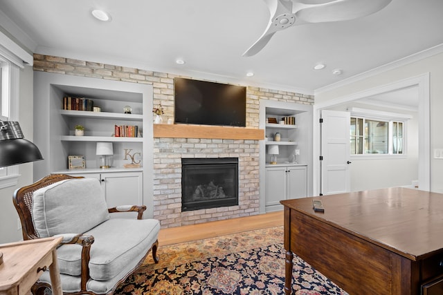 living room featuring built in shelves, a brick fireplace, light hardwood / wood-style flooring, ornamental molding, and ceiling fan