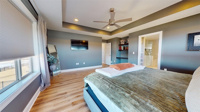 bedroom with light hardwood / wood-style flooring, a tray ceiling, ensuite bath, and ceiling fan