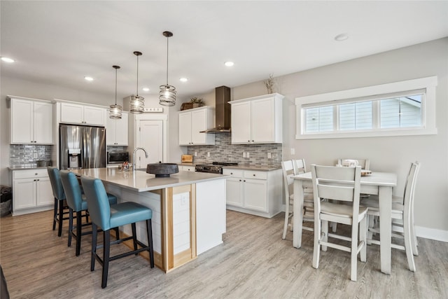 kitchen with pendant lighting, appliances with stainless steel finishes, a kitchen island with sink, white cabinets, and wall chimney exhaust hood