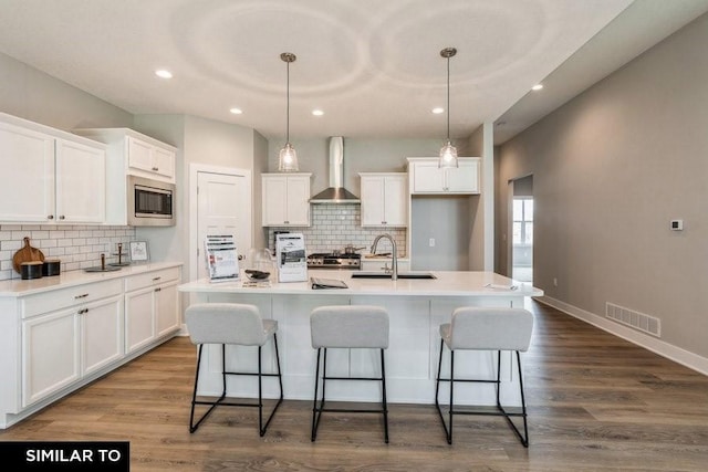 kitchen featuring sink, wall chimney range hood, a kitchen island with sink, white cabinetry, and stainless steel microwave