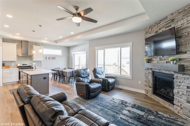 living room featuring sink, a tray ceiling, ceiling fan, a fireplace, and light hardwood / wood-style floors