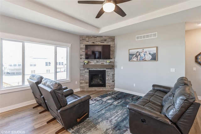 living room featuring a tray ceiling, a stone fireplace, wood-type flooring, and ceiling fan