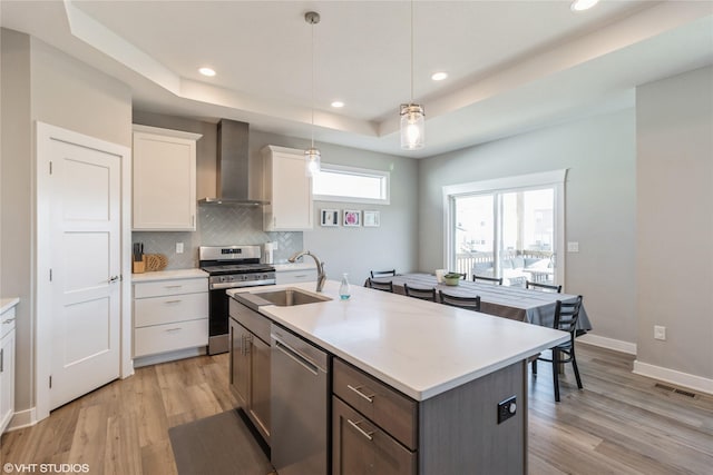 kitchen with stainless steel appliances, white cabinets, decorative light fixtures, a raised ceiling, and wall chimney exhaust hood