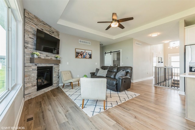 1210 NW Pine Bluff Cir, Waukee, IA 50263 - living room featuring light wood-type flooring, a stone fireplace, a ceiling fan, a tray ceiling, and a barn door