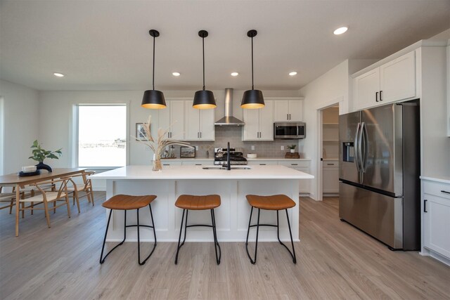 kitchen with stainless steel appliances, hanging light fixtures, a kitchen island with sink, and wall chimney range hood