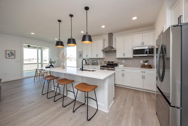 kitchen featuring wall chimney range hood, sink, appliances with stainless steel finishes, a kitchen island with sink, and hanging light fixtures