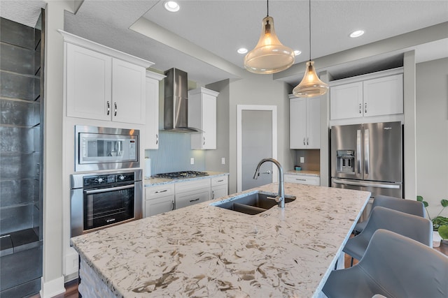 kitchen with white cabinetry, an island with sink, sink, stainless steel appliances, and wall chimney range hood