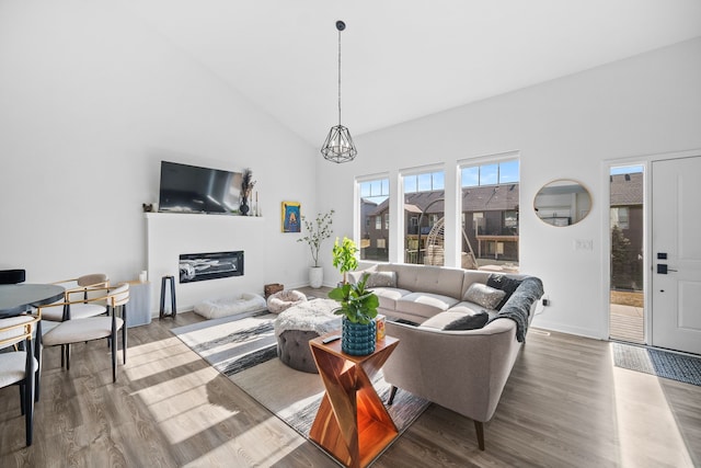 living room featuring wood finished floors, baseboards, high vaulted ceiling, an inviting chandelier, and a glass covered fireplace