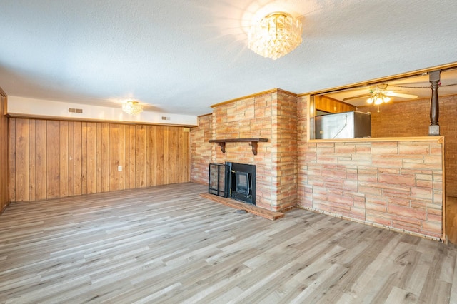 unfurnished living room with ceiling fan, a fireplace, a textured ceiling, light wood-type flooring, and wood walls