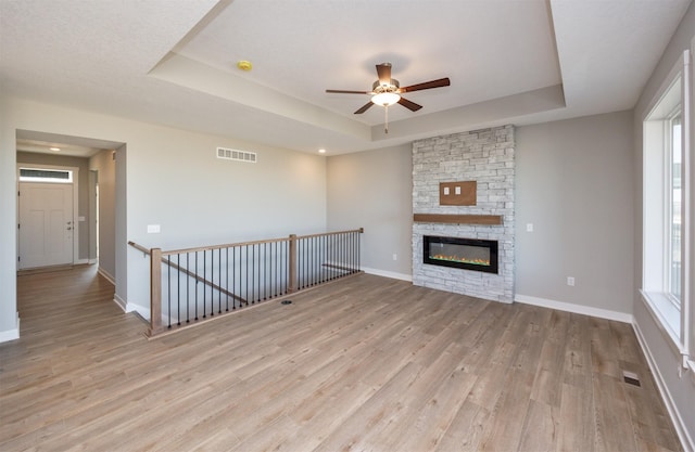 unfurnished living room with a stone fireplace, light hardwood / wood-style floors, a raised ceiling, and ceiling fan