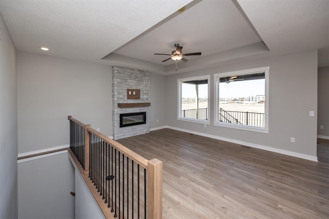 unfurnished living room with hardwood / wood-style flooring, a stone fireplace, ceiling fan, and a tray ceiling