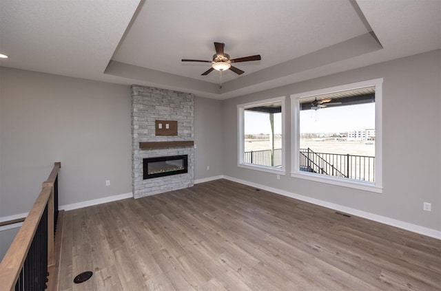 unfurnished living room with ceiling fan, a stone fireplace, light hardwood / wood-style floors, and a tray ceiling