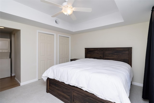 bedroom featuring two closets, stacked washer / dryer, ceiling fan, a tray ceiling, and light carpet