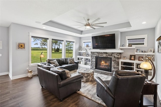 living room with a stone fireplace, dark hardwood / wood-style floors, a raised ceiling, and ceiling fan