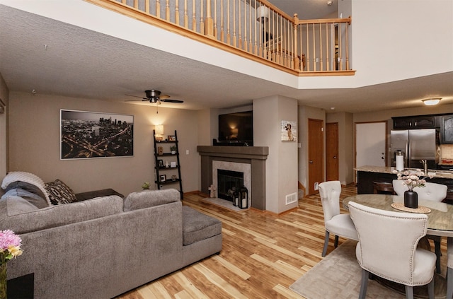 living room featuring a fireplace, a high ceiling, ceiling fan, a textured ceiling, and light wood-type flooring