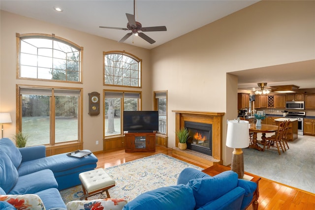 living room featuring a glass covered fireplace, a ceiling fan, a high ceiling, and light wood-style floors