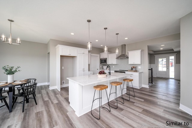 kitchen with pendant lighting, an island with sink, white cabinets, wall chimney exhaust hood, and light wood-type flooring