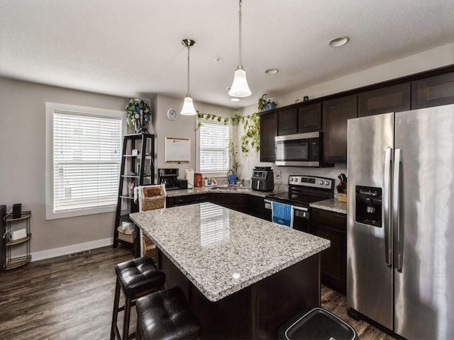 kitchen featuring appliances with stainless steel finishes, hanging light fixtures, a center island, light stone countertops, and dark brown cabinets