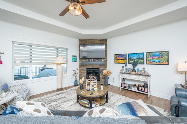 living room with a tray ceiling, wood-type flooring, a stone fireplace, and ceiling fan