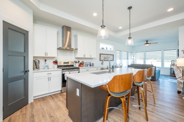 kitchen with sink, wall chimney range hood, stainless steel gas stove, white cabinetry, and a raised ceiling