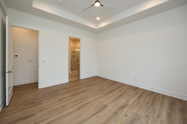 unfurnished bedroom featuring ceiling fan, ensuite bath, light wood-type flooring, and a tray ceiling