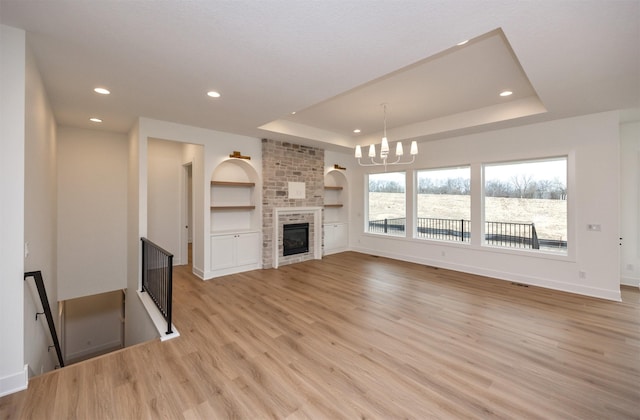 unfurnished living room with built in shelves, a chandelier, light wood-type flooring, a tray ceiling, and a fireplace