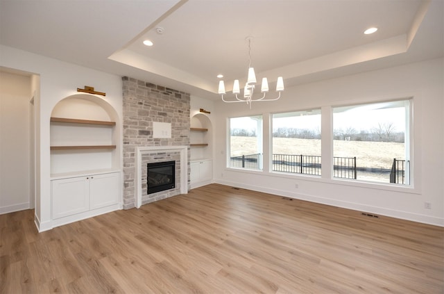 unfurnished living room featuring built in features, a large fireplace, a chandelier, a tray ceiling, and light hardwood / wood-style flooring