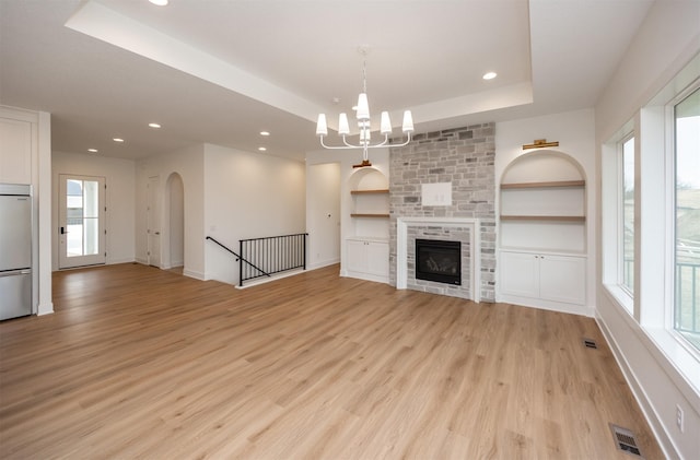 unfurnished living room with an inviting chandelier, a tray ceiling, a fireplace, light hardwood / wood-style floors, and built in shelves