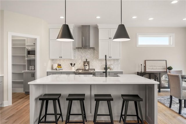 kitchen featuring white cabinets, hanging light fixtures, a kitchen island with sink, stainless steel appliances, and wall chimney range hood