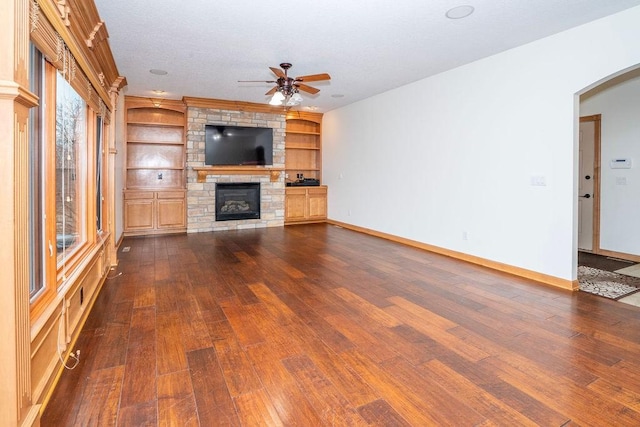 unfurnished living room with built in shelves, ceiling fan, a stone fireplace, and dark hardwood / wood-style flooring