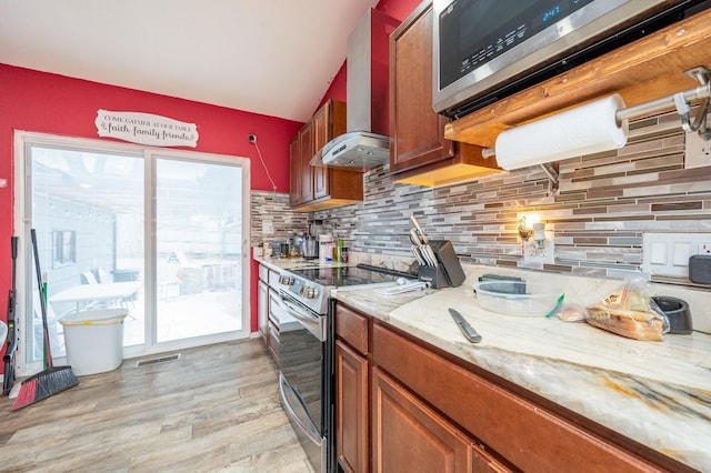 kitchen with wall chimney range hood, light hardwood / wood-style flooring, appliances with stainless steel finishes, light stone counters, and decorative backsplash