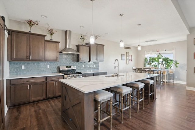 kitchen with decorative light fixtures, stainless steel gas stove, a kitchen breakfast bar, wall chimney range hood, and a center island with sink