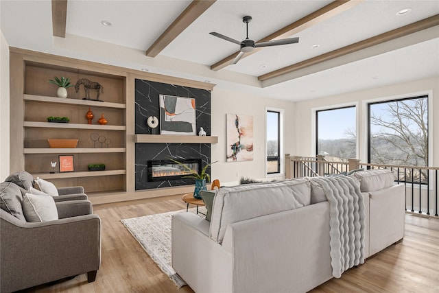 living room with built in shelves, light hardwood / wood-style flooring, beamed ceiling, ceiling fan, and a premium fireplace