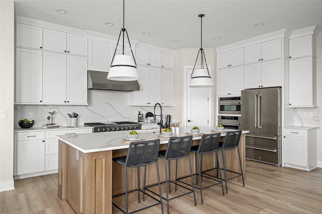 kitchen featuring wall chimney range hood, high end fridge, a kitchen island with sink, hanging light fixtures, and white cabinets