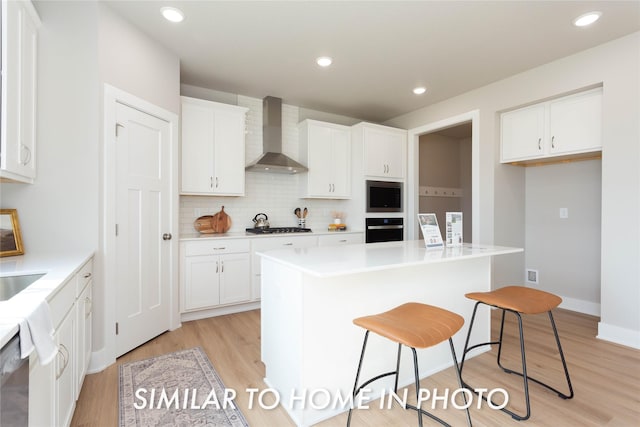kitchen with stainless steel appliances, wall chimney exhaust hood, light wood finished floors, white cabinetry, and a center island