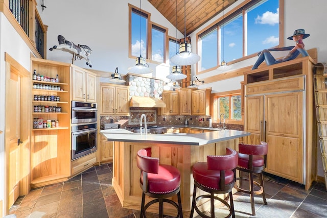 kitchen with a breakfast bar, an island with sink, backsplash, hanging light fixtures, and stainless steel double oven