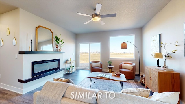 living area featuring baseboards, a ceiling fan, a glass covered fireplace, wood finished floors, and a textured ceiling