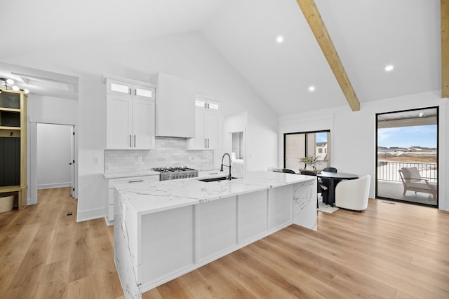 kitchen featuring a kitchen island with sink, a sink, light stone counters, white cabinetry, and decorative backsplash