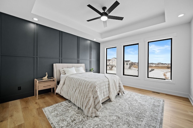 bedroom with baseboards, light wood-style flooring, recessed lighting, a raised ceiling, and a decorative wall