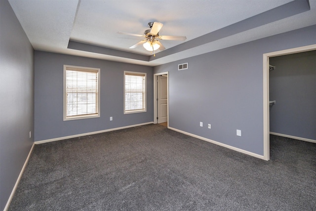 unfurnished bedroom with dark carpet, ceiling fan, a walk in closet, and a tray ceiling