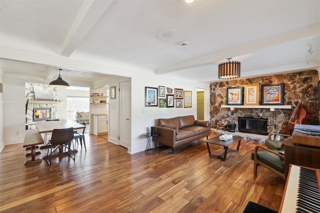 living room with a textured ceiling, a stone fireplace, wood finished floors, beamed ceiling, and baseboards