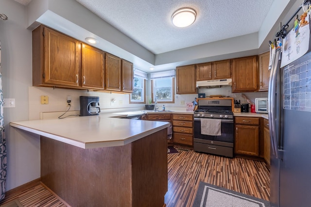 kitchen featuring light countertops, appliances with stainless steel finishes, brown cabinetry, a peninsula, and under cabinet range hood