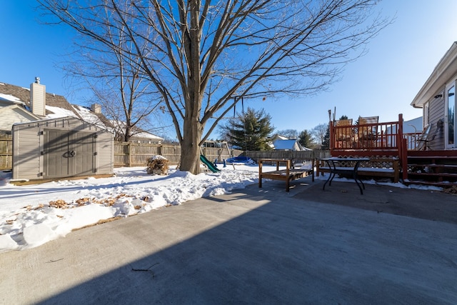 yard layered in snow with a storage shed, a fenced backyard, a deck, an outdoor structure, and a playground