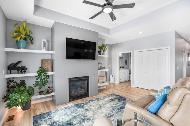 living room with light wood finished floors, visible vents, a glass covered fireplace, ceiling fan, and a tray ceiling