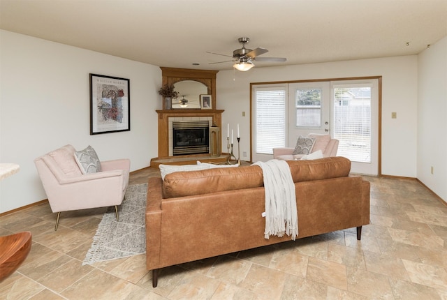 living room featuring a glass covered fireplace, stone finish flooring, ceiling fan, and baseboards