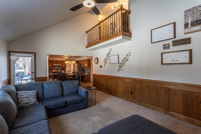 living area featuring wooden walls, light colored carpet, a wainscoted wall, high vaulted ceiling, and ceiling fan with notable chandelier