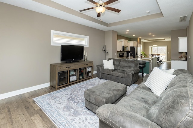 living area with a raised ceiling, visible vents, a ceiling fan, wood finished floors, and baseboards